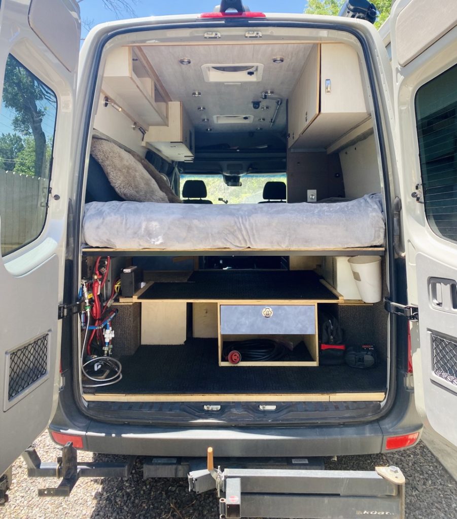 A 4x4 Mercedes-Benz Sprinter camper van is parked with the rear doors open, revealing a platform bed with storage underneath.